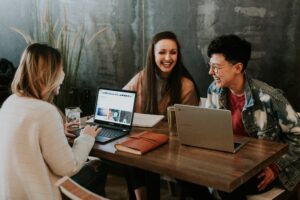 Three women sitting at a table laughing in front of computers